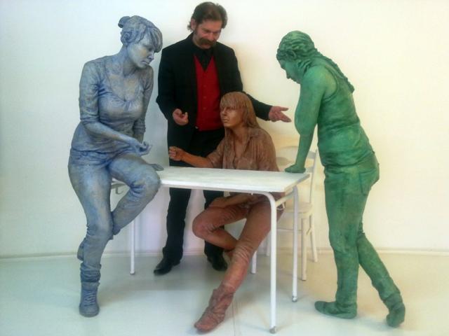 Instalacja artystyczna przedstawiająca trzy postacie kobiece w różnych odcieniach (niebieskim, brązowym i zielonym) oraz mężczyznę w czarnym garniturze i czerwonej kamizelce, stojących wokół...