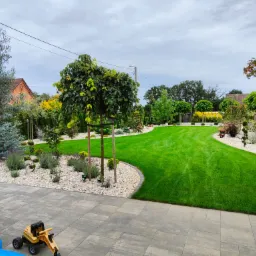 Zadbany ogród z równo przystrzyżonym trawnikiem, ozdobnymi drzewami i krzewami, wyłożony kamieniami, z kostką brukową na pierwszym planie i zabawkami dziecięcymi.