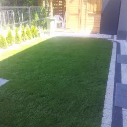 Świeżo założony trawnik z rolki, drewniana altana w tle, obrzeża z kostki brukowej, młode tuje wzdłuż ogrodzenia.