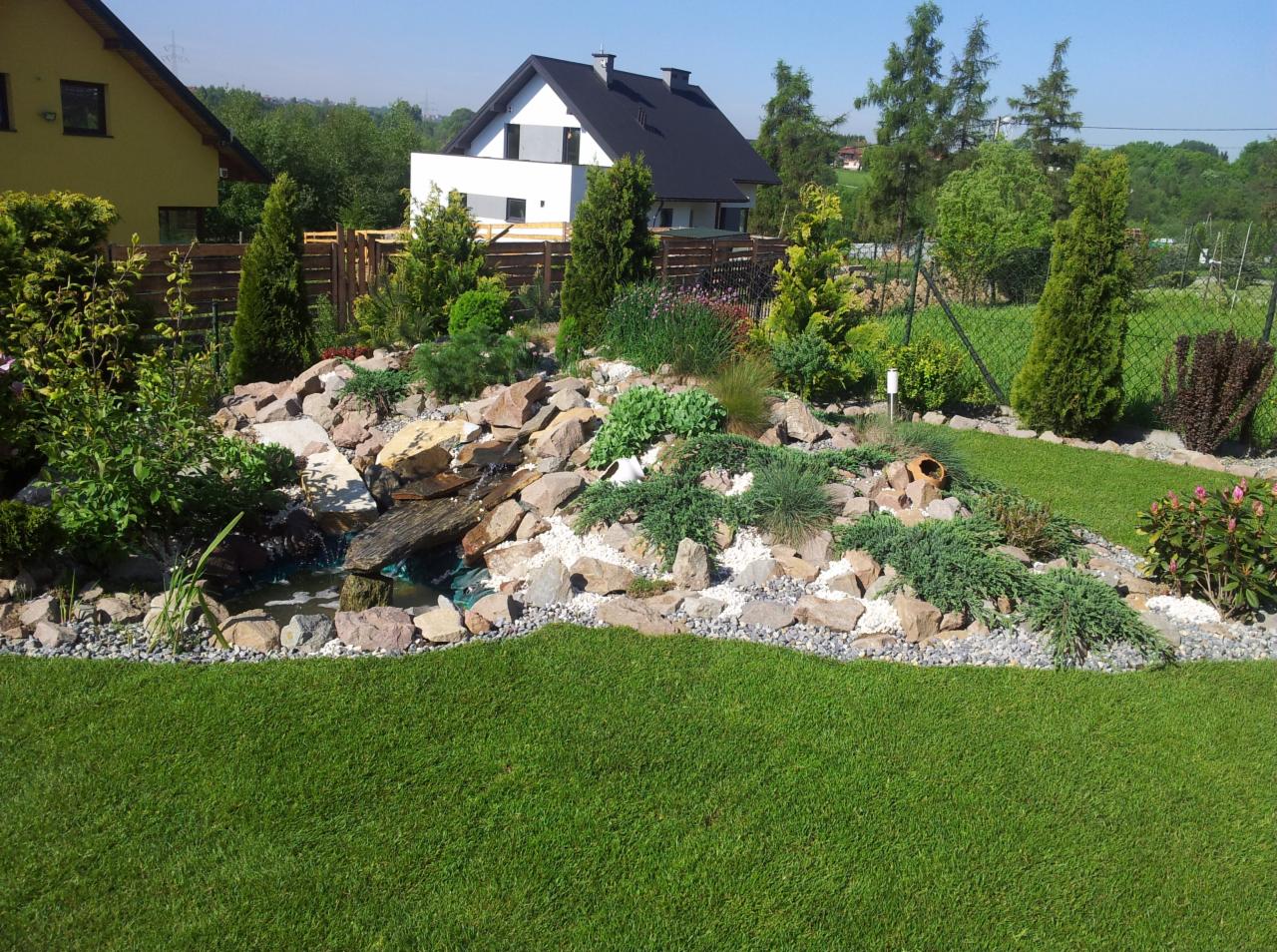 Ogród z kaskadą wodną, kamieniami i roślinami ozdobnymi na tle domu. Zieleń trawnika kontrastuje z jasnymi kamieniami. Zadbany trawnik, dobrze oświetlony.