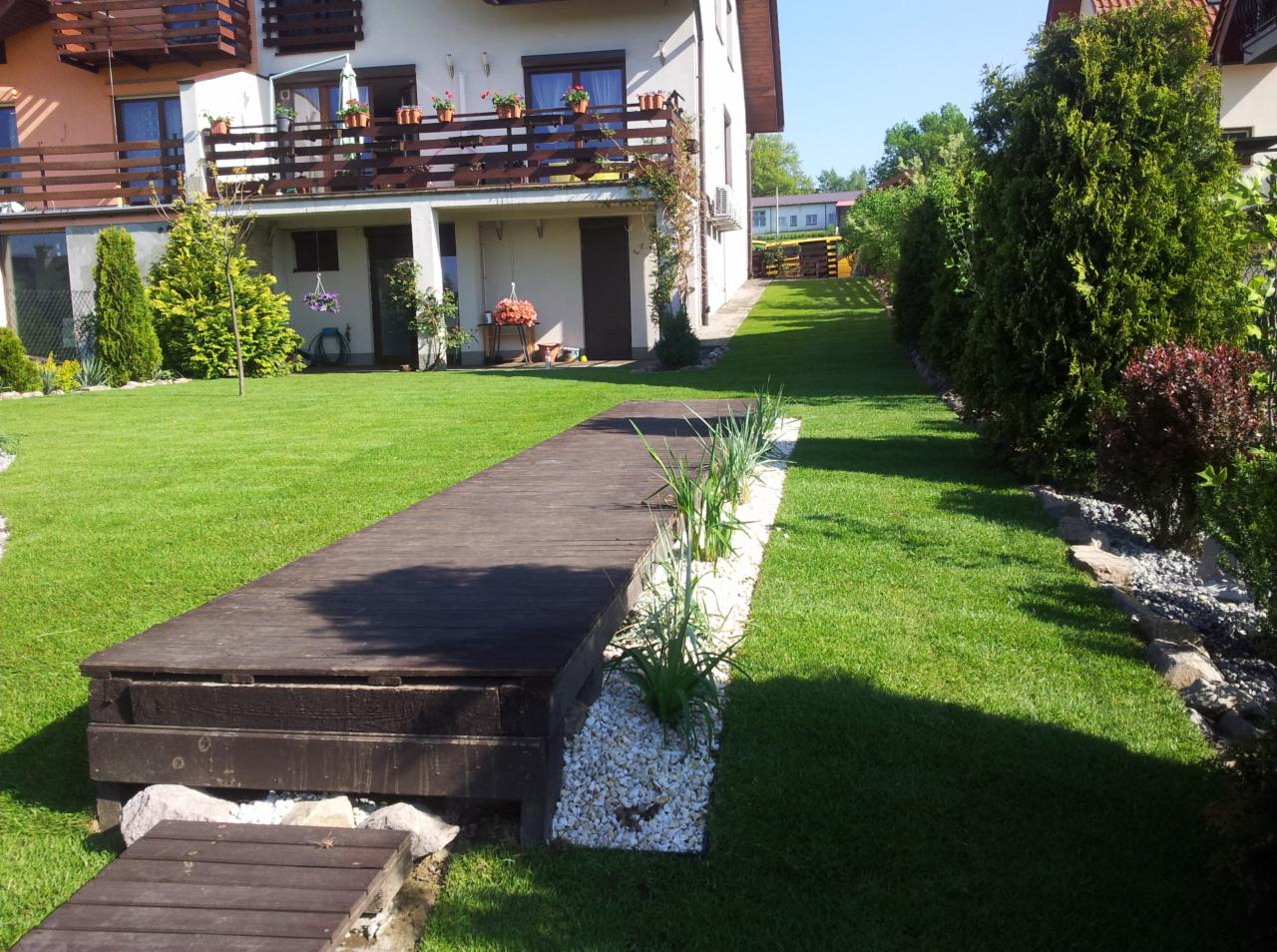 Zadbany ogród z równo przystrzyżonym trawnikiem, drewnianym podestem i rabatami z białych kamieni. W tle widoczny dom z balkonami i zielenią.