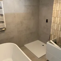 Nowoczesna łazienka w trakcie wykańczania: biała wanna wolnostojąca, brodzik, grzejnik drabinkowy i częściowo zamontowana szafka pod umywalkę.