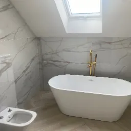 Nowoczesna łazienka na poddaszu z białą wanną wolnostojącą i marmurowymi płytkami. Złota armatura, okno dachowe. Jasne, przestronne wnętrze.