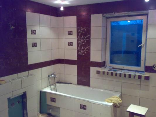 Wnętrze łazienki w trakcie remontu, widoczne białe i bordowe płytki, wanna obudowana płytkami, okno z niebieską folią ochronną oraz fragment ściany z przygotowanym otworem pod zabudowę.