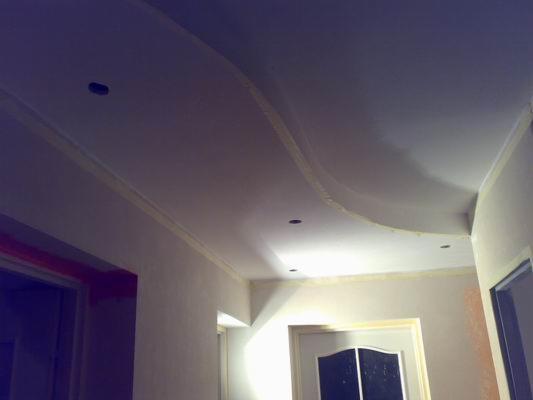 Biały sufit podwieszany z ozdobnym, falistym obniżeniem i zamontowanymi punktowymi źródłami światła, widoczne fragmenty ścian i drzwi.