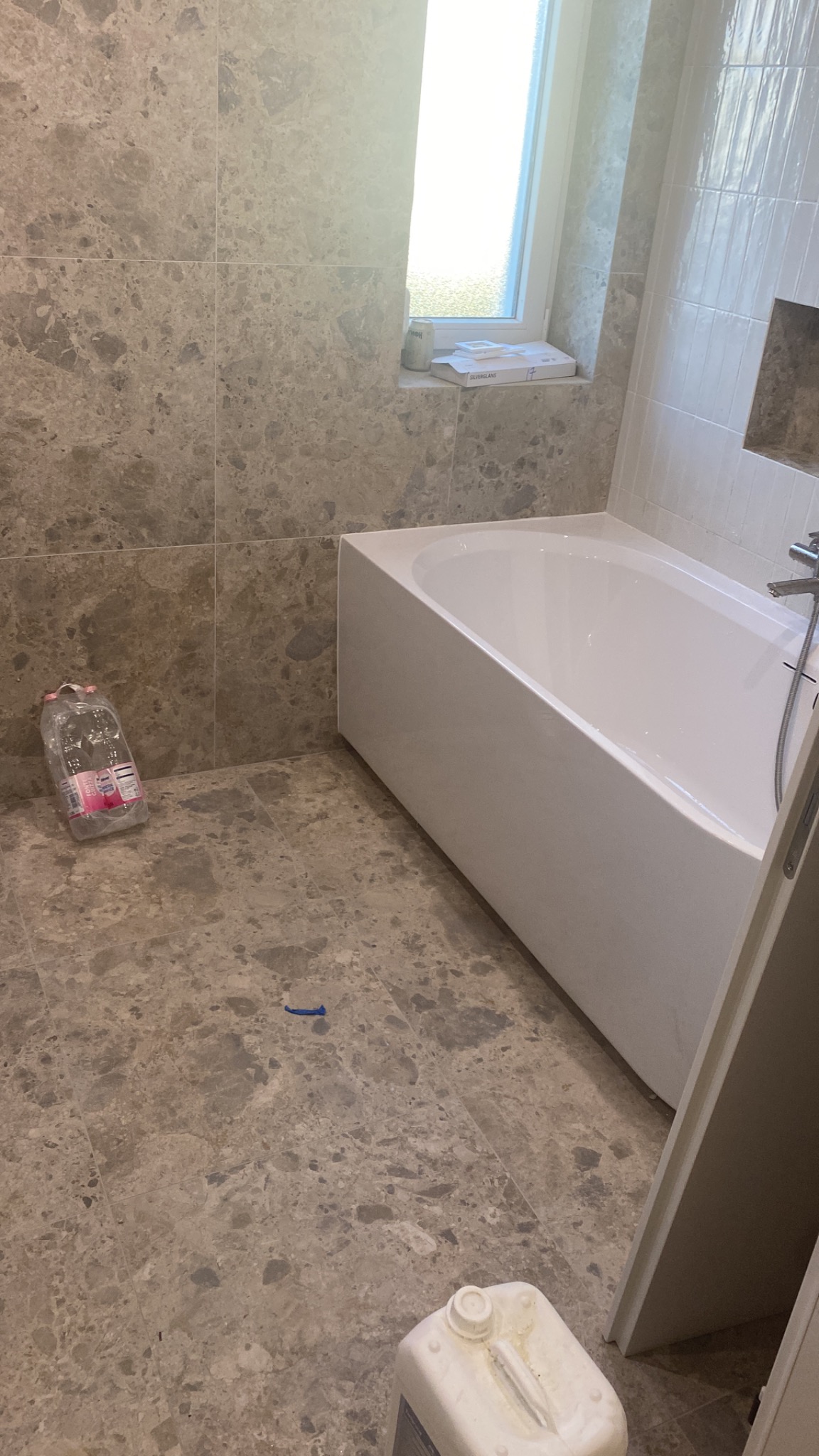 Wykończona łazienka z wanną, marmurowymi płytkami na podłodze i ścianach. Widoczne akcesoria do czyszczenia. Jasne światło wpada przez okno.