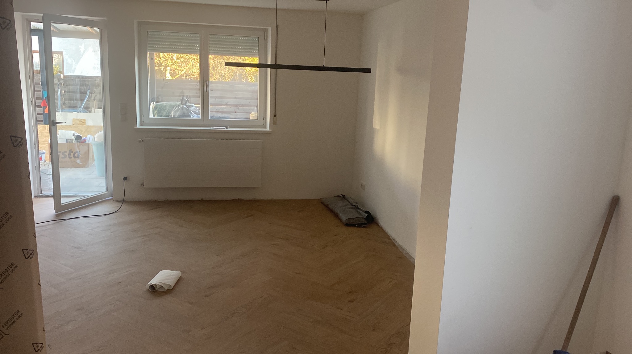 Jasne, parkietowe podłogi ułożone w jodełkę w pustym pokoju z oknem i grzejnikiem. Ściany białe, częściowo zabezpieczone. Widoczne narzędzia i materiały budowlane.