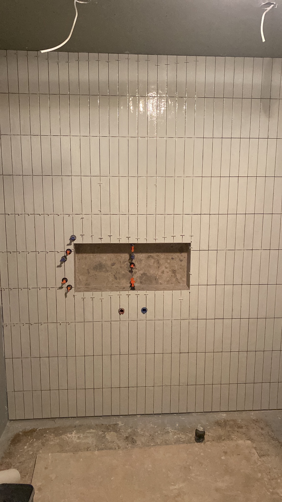 Ściana łazienki w trakcie układania podłużnych, jasnych płytek z widocznym otworem na półkę i wystającymi elementami instalacji wodnej.