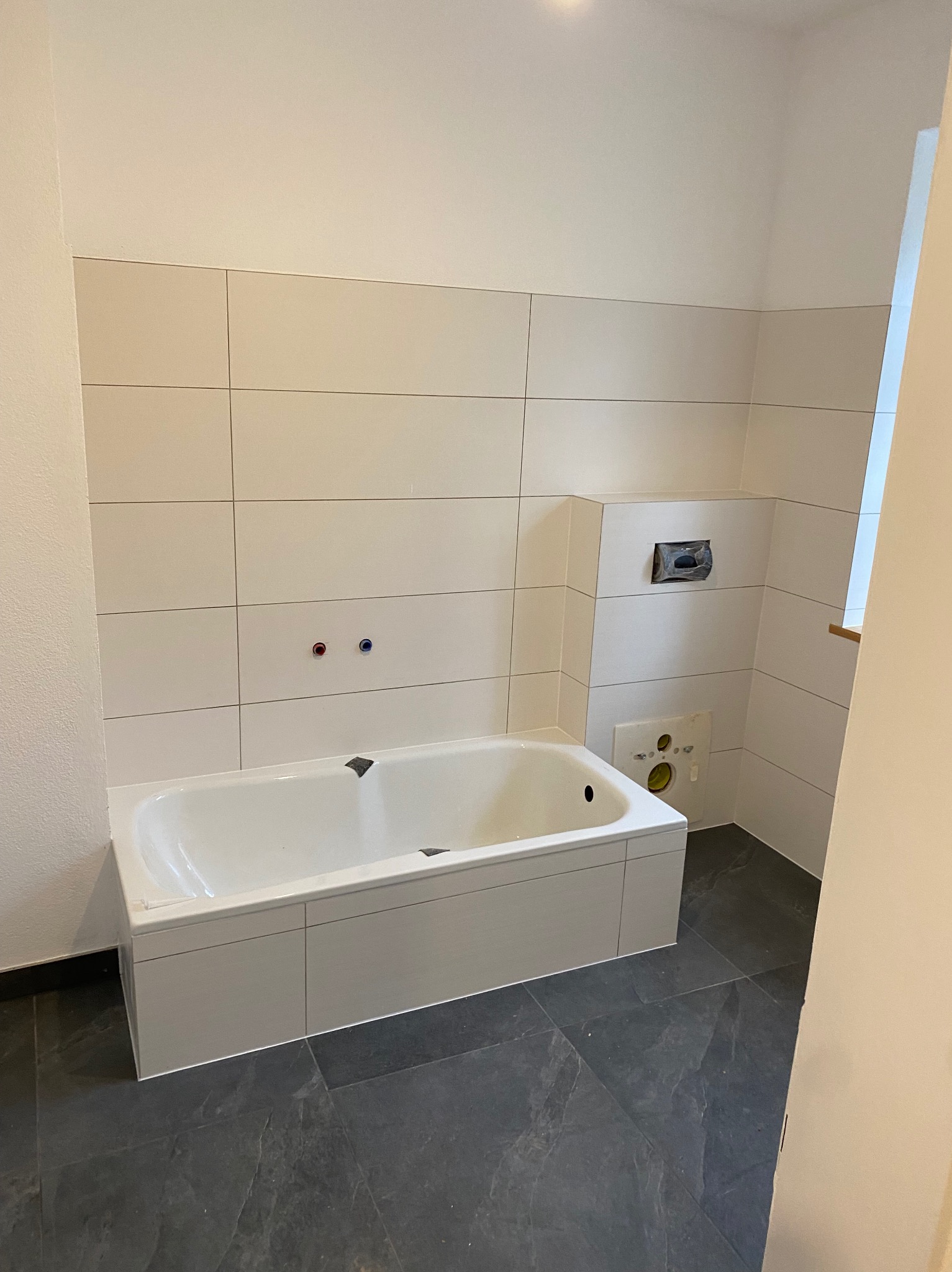 Wykończona łazienka z wanną obudowaną płytkami, kremowe płytki ścienne, ciemnoszara podłoga, widoczne przyłącza wody i instalacja podtynkowa WC.