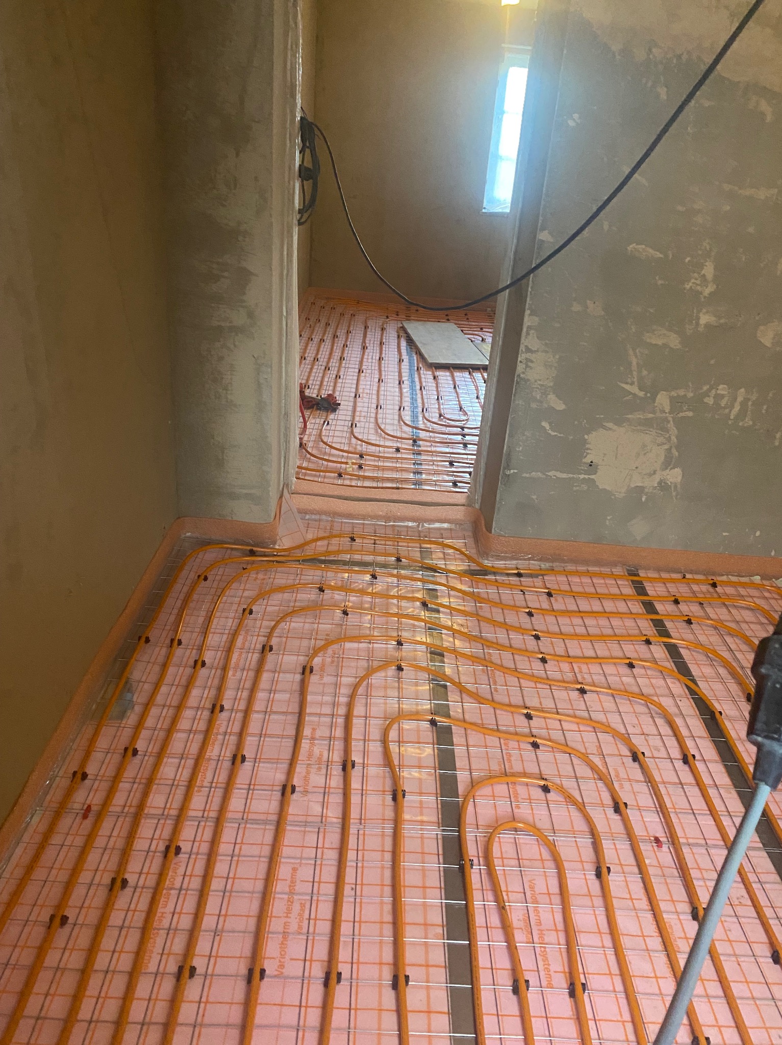 Pomarańczowe rurki ogrzewania podłogowego ułożone na różowej macie, przymocowane do metalowej siatki, widoczne na tle surowych ścian w trakcie remontu.