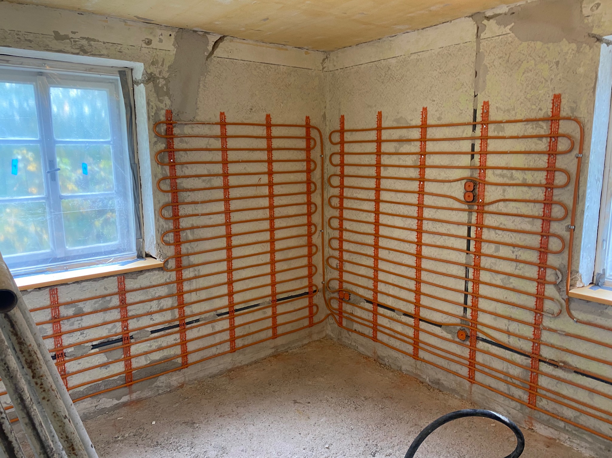 Pomieszczenie w trakcie montażu ogrzewania podłogowego ściennego. Widoczne rury grzewcze w kolorze pomarańczowym, przymocowane do surowych ścian. Okna zabezpieczone folią.