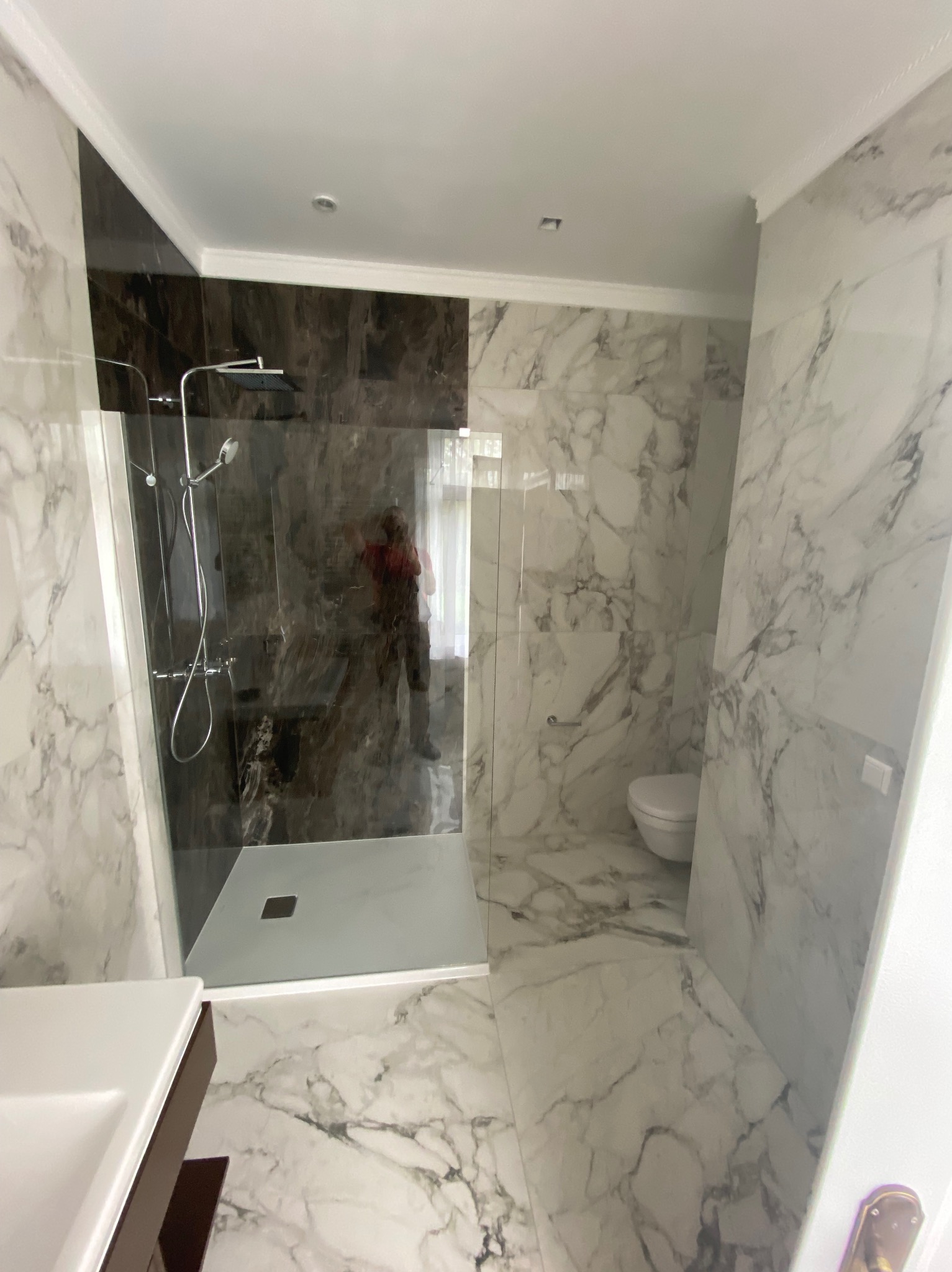 Wykończona łazienka z kabiną typu walk-in, marmurowymi płytkami na ścianach i podłodze, oraz toaletą w rogu. Widoczna armatura w odcieniach srebra i brązu.