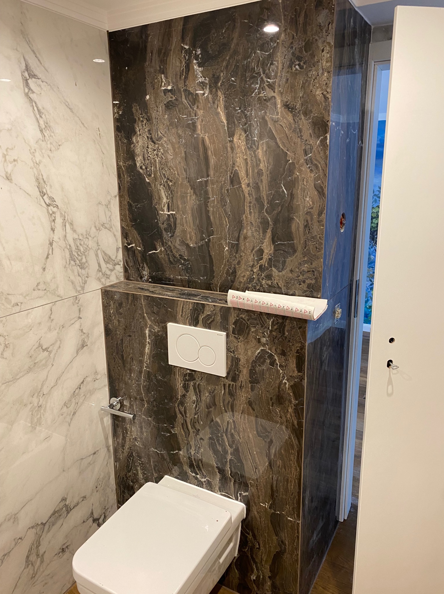 Nowoczesna toaleta podwieszana z brązowymi płytkami imitującymi marmur i białymi płytkami na ścianie. Widoczny przycisk spłuczki i częściowo rozwinięta miarka.