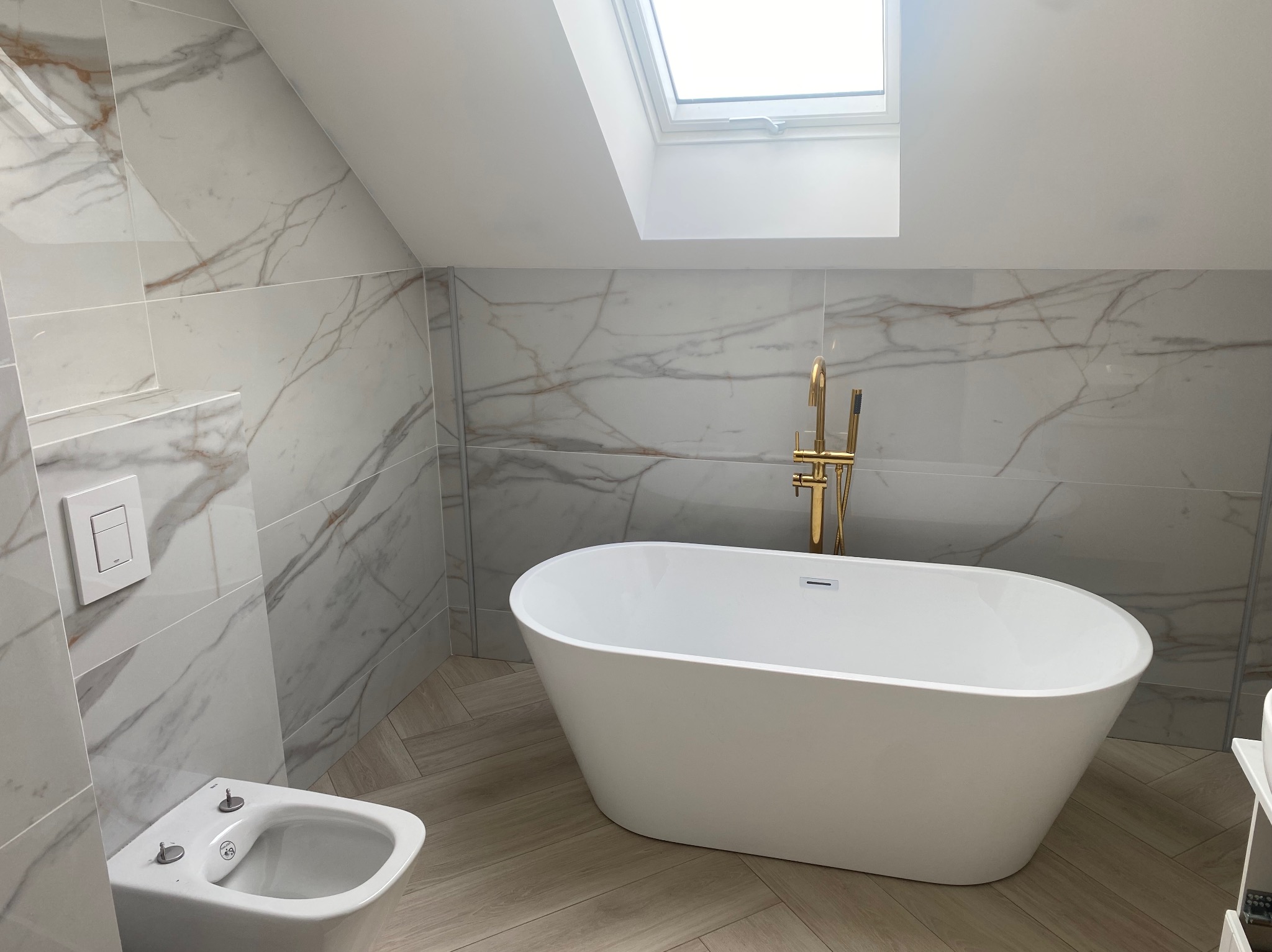 Nowoczesna łazienka na poddaszu z białą wanną wolnostojącą i marmurowymi płytkami. Złota armatura, okno dachowe. Jasne, przestronne wnętrze.