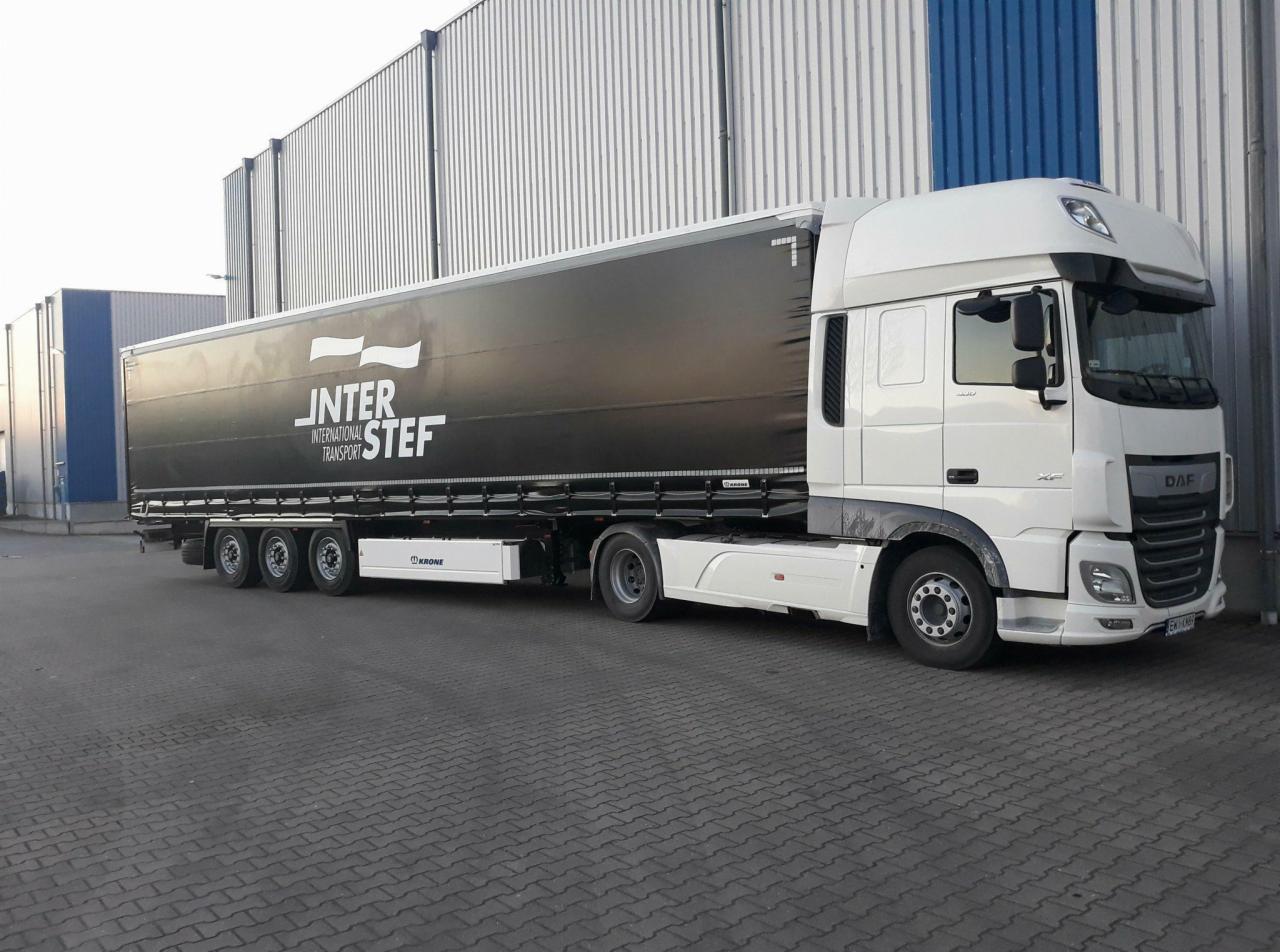 Biała ciężarówka DAF XF z czarną naczepą firmy Inter Stef International Transport na tle szarego magazynu z niebieskim akcentem.