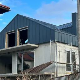 Surowy stan domu w trakcie budowy z zamontowaną, ciemnoszarą blachą na dachu i przygotowanymi otworami okiennymi oraz rusztowaniami.