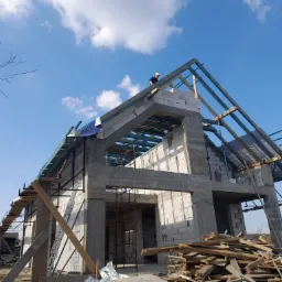 Wznoszenie dwukondygnacyjnego domu z betonu komórkowego, widoczna konstrukcja dachu z drewnianych belek, pracownik na szczycie, niebieskie niebo z chmurami w tle.