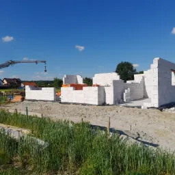 Wznoszenie ścian parteru domu z białych bloczków na fundamencie, widoczny dźwig budowlany i teren budowy.