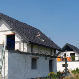 Montaż czarnej dachówki na nowym, murowanym domu jednorodzinnym, widoczny dekarz przy pracy, rusztowanie na pierwszym piętrze, stan surowy otwarty.