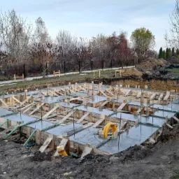 Zalane betonem szalunki fundamentów domu jednorodzinnego, widoczne żółte rury instalacyjne, w tle wykop ziemny, drzewa i zabudowa jednorodzinna.