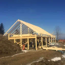 Wznoszenie konstrukcji domu szkieletowego z drewnianą więźbą dachową na tle błękitnego nieba i zimowego krajobrazu.