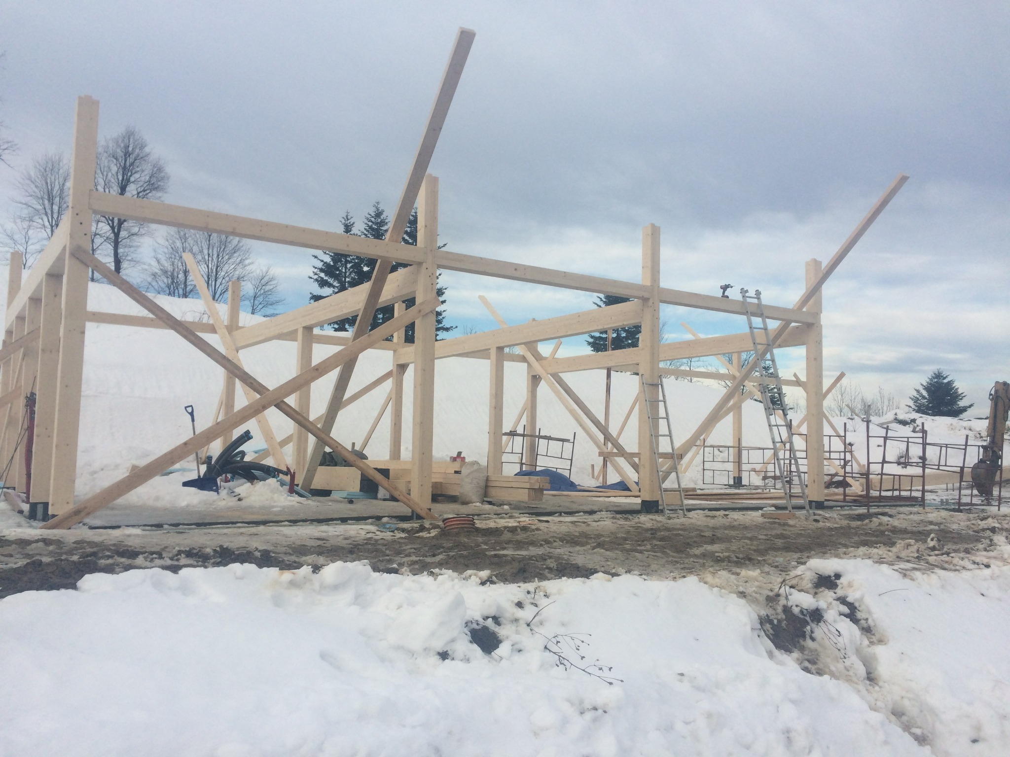 Wznoszenie szkieletu drewnianego domu w zimowej scenerii, widoczne rusztowania, drabina i częściowo odśnieżony teren budowy.