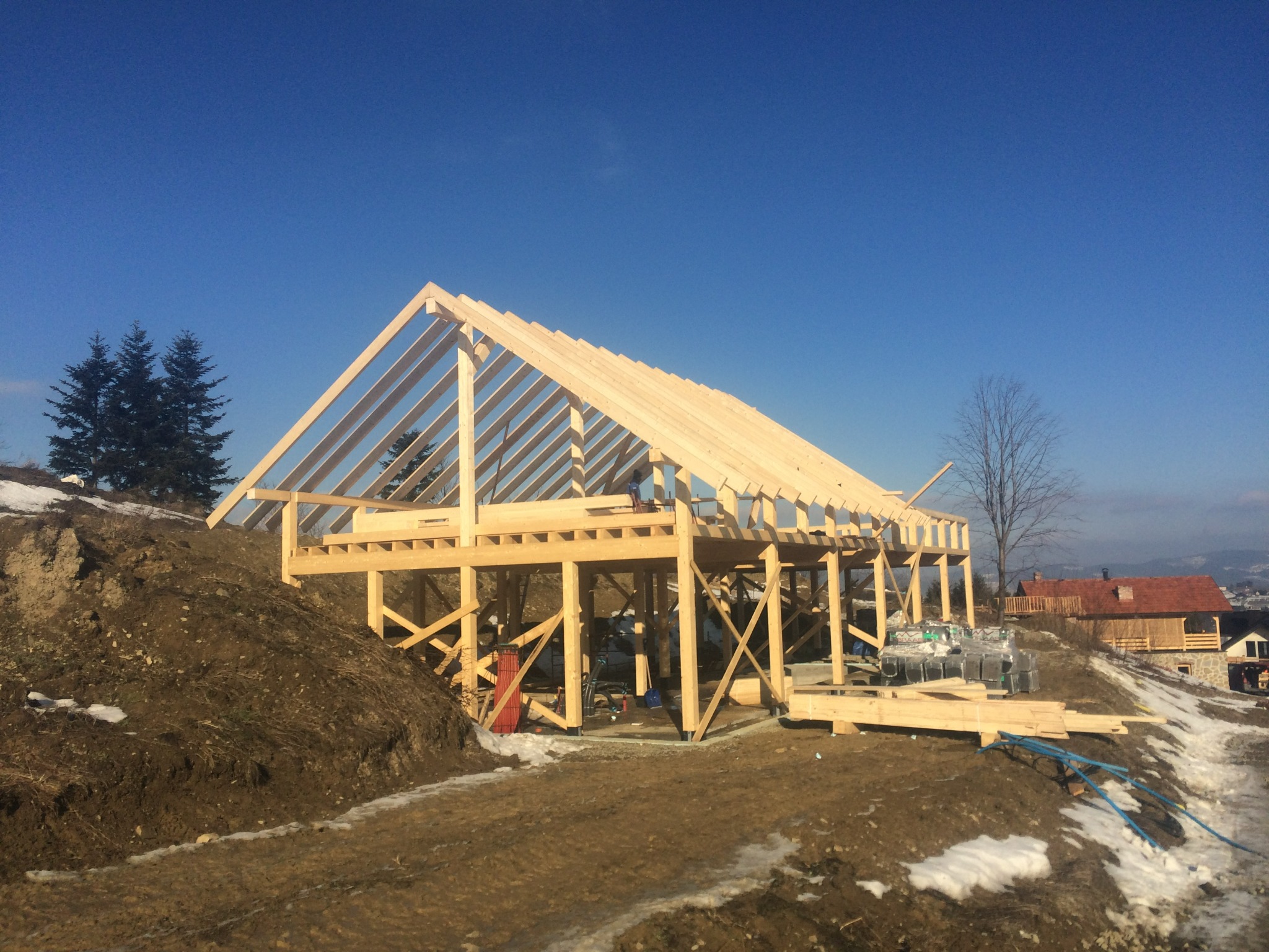 Wznoszenie konstrukcji domu szkieletowego z drewnianą więźbą dachową na tle błękitnego nieba i zimowego krajobrazu.
