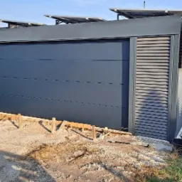 Nowoczesny garaż z szarą bramą segmentową i panelami słonecznymi na dachu, widoczne szalunki fundamentowe w trakcie budowy, w tle domy jednorodzinne i plac zabaw.