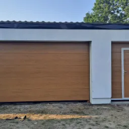 Nowoczesny garaż z segmentową bramą garażową w kolorze jasnego drewna i dopasowanymi drzwiami bocznymi z aluminiową ramą, na tle białej elewacji budynku.