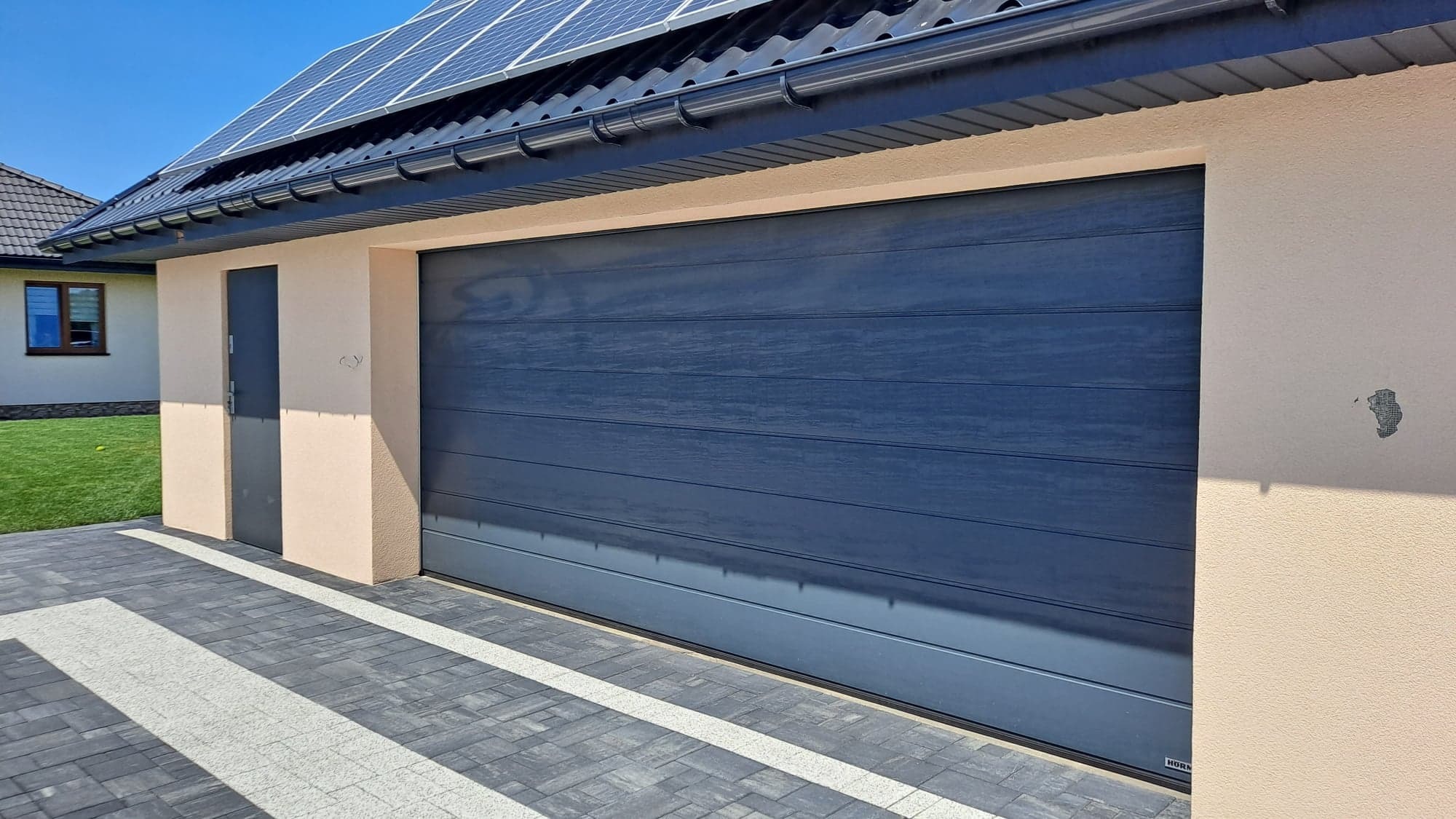 Nowoczesna, panelowa brama garażowa w kolorze grafitowym, zamontowana w budynku z panelami słonecznymi na dachu, Konin. Widoczny fragment chodnika z kostki brukowej.