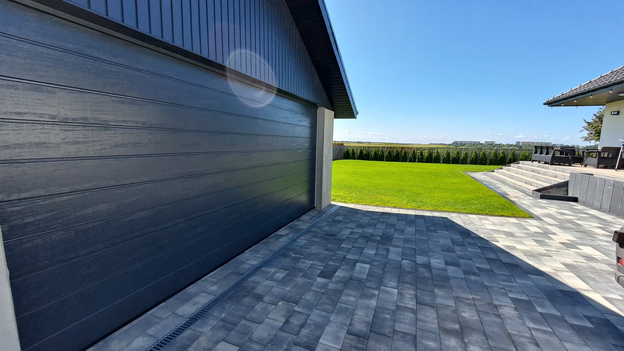 Nowoczesna, segmentowa brama garażowa w kolorze antracytowym z poziomymi przetłoczeniami, widok z zewnątrz na tle zadbanego trawnika i domu z szarej kostki brukowej.