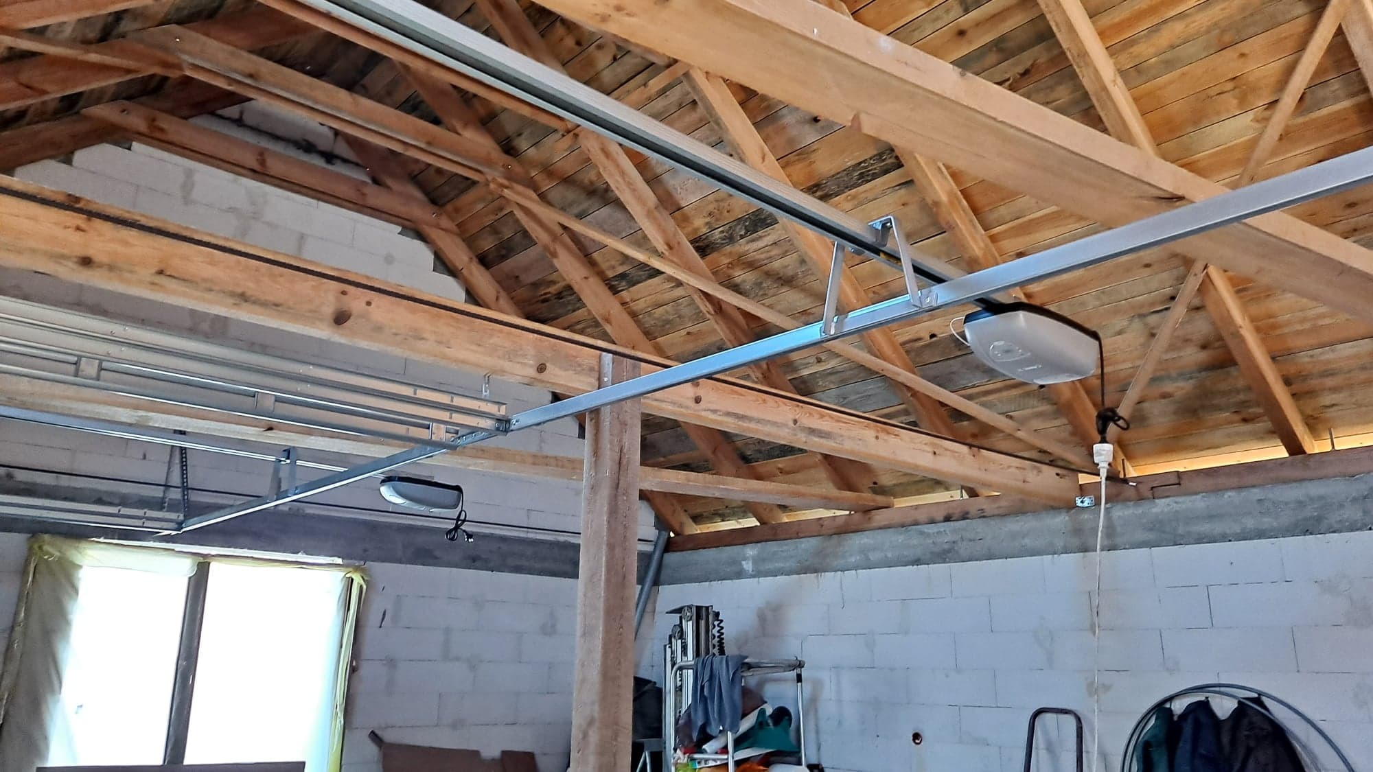 Montaż szyny i napędu elektrycznego do bramy garażowej, widok na surowy sufit z drewnianą konstrukcją i białe ściany z bloczków.