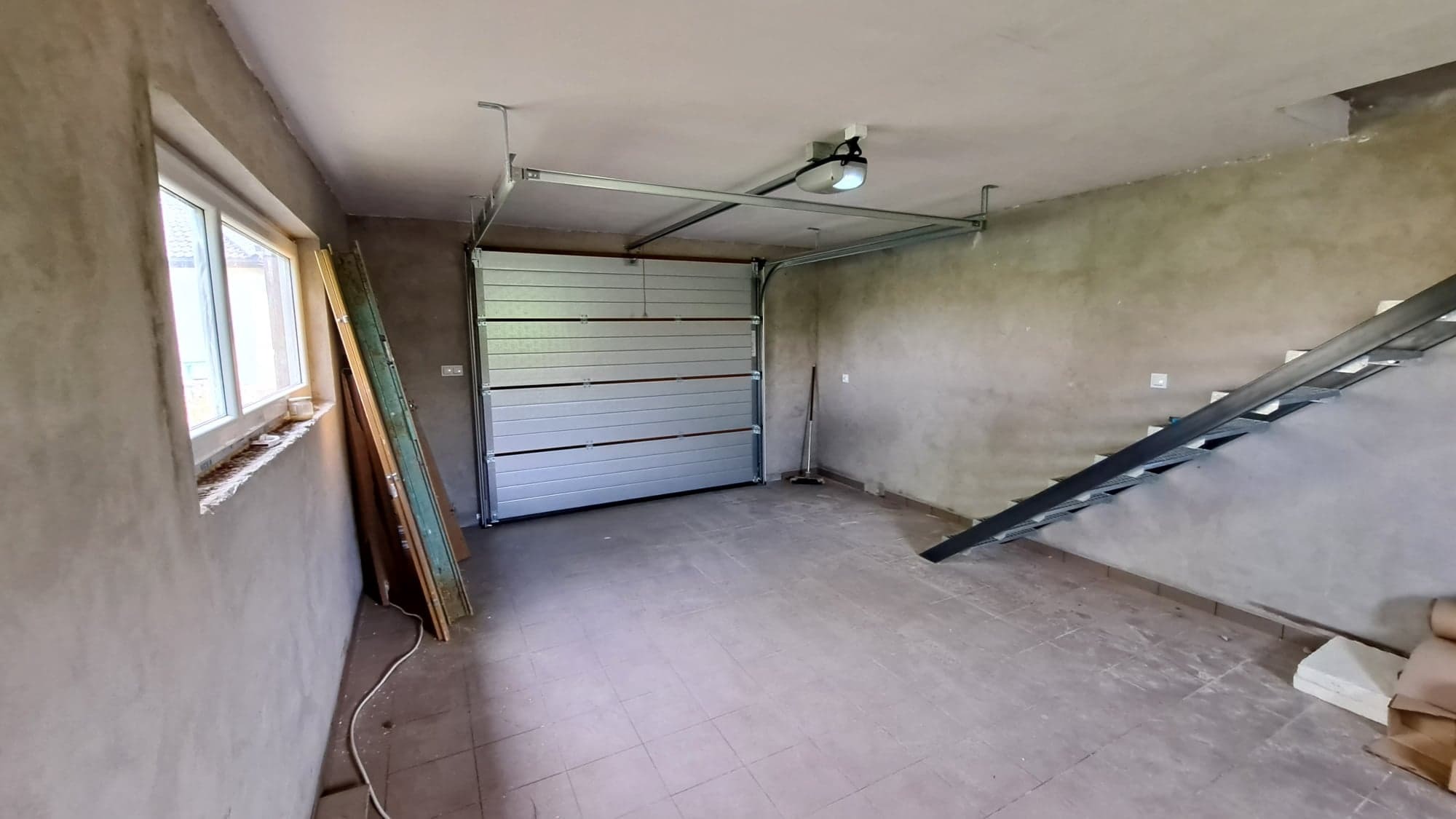 Wnętrze garażu z zamontowaną segmentową bramą garażową w kolorze jasnoszarym, widoczny mechanizm podnoszenia, surowe ściany i podłoga z płytek, częściowo widoczne schody metalowe i okno.