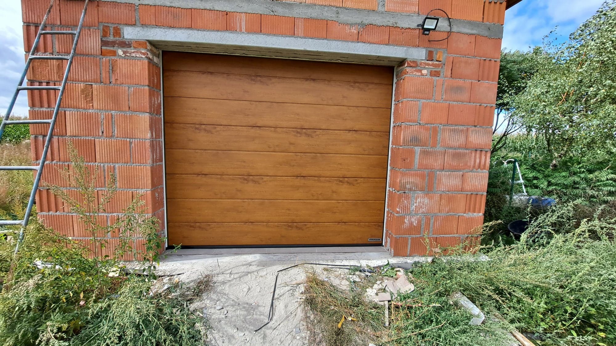 Zamknięta, segmentowa brama garażowa w kolorze jasnego drewna, zamontowana w surowym murze z czerwonej cegły, z widoczną drabinką z boku i roślinnością u podstawy.