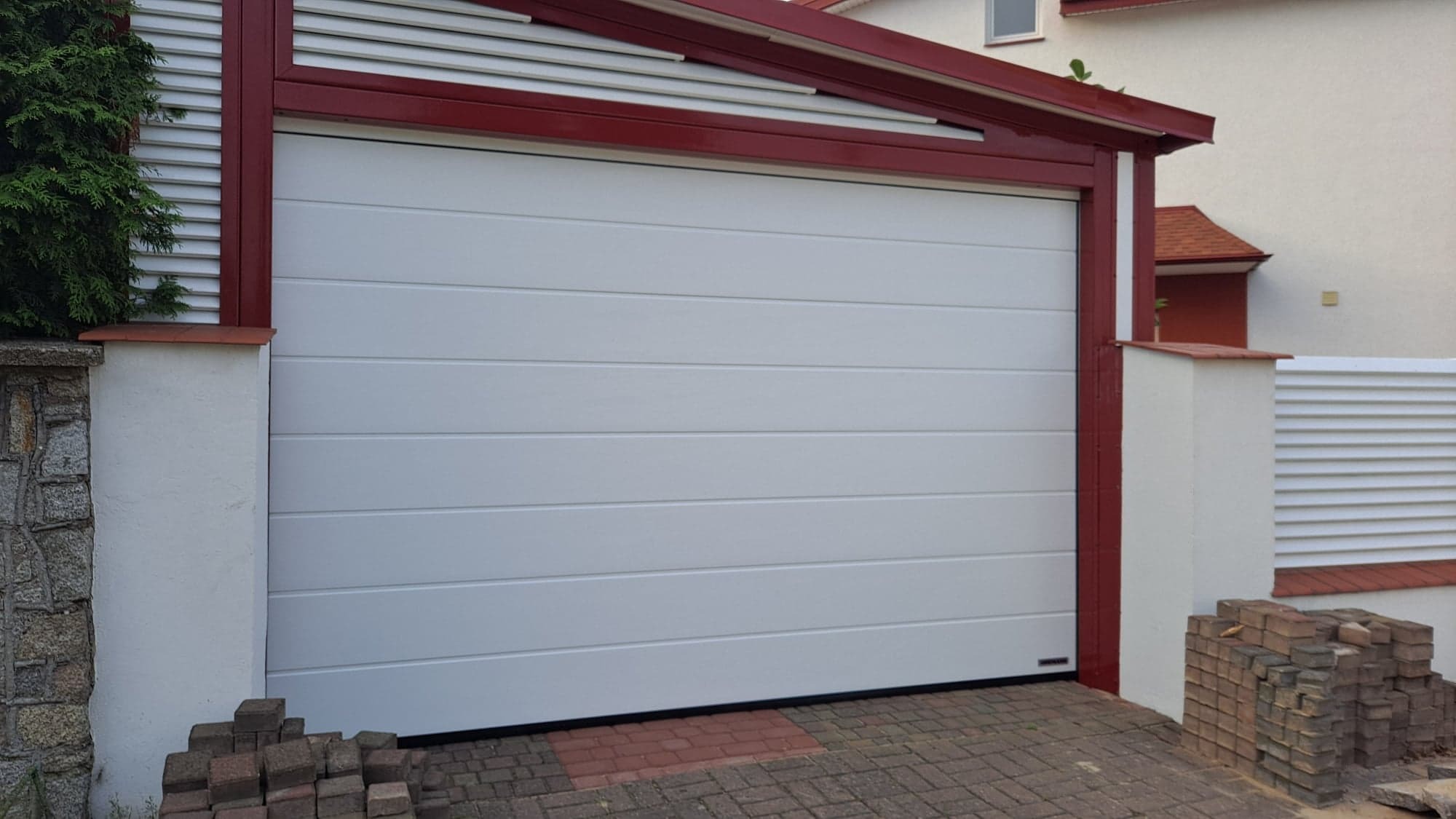 Biała segmentowa brama garażowa z czerwoną ramą, widoczna od zewnątrz, z kostką brukową i stertą cegieł u podstawy.