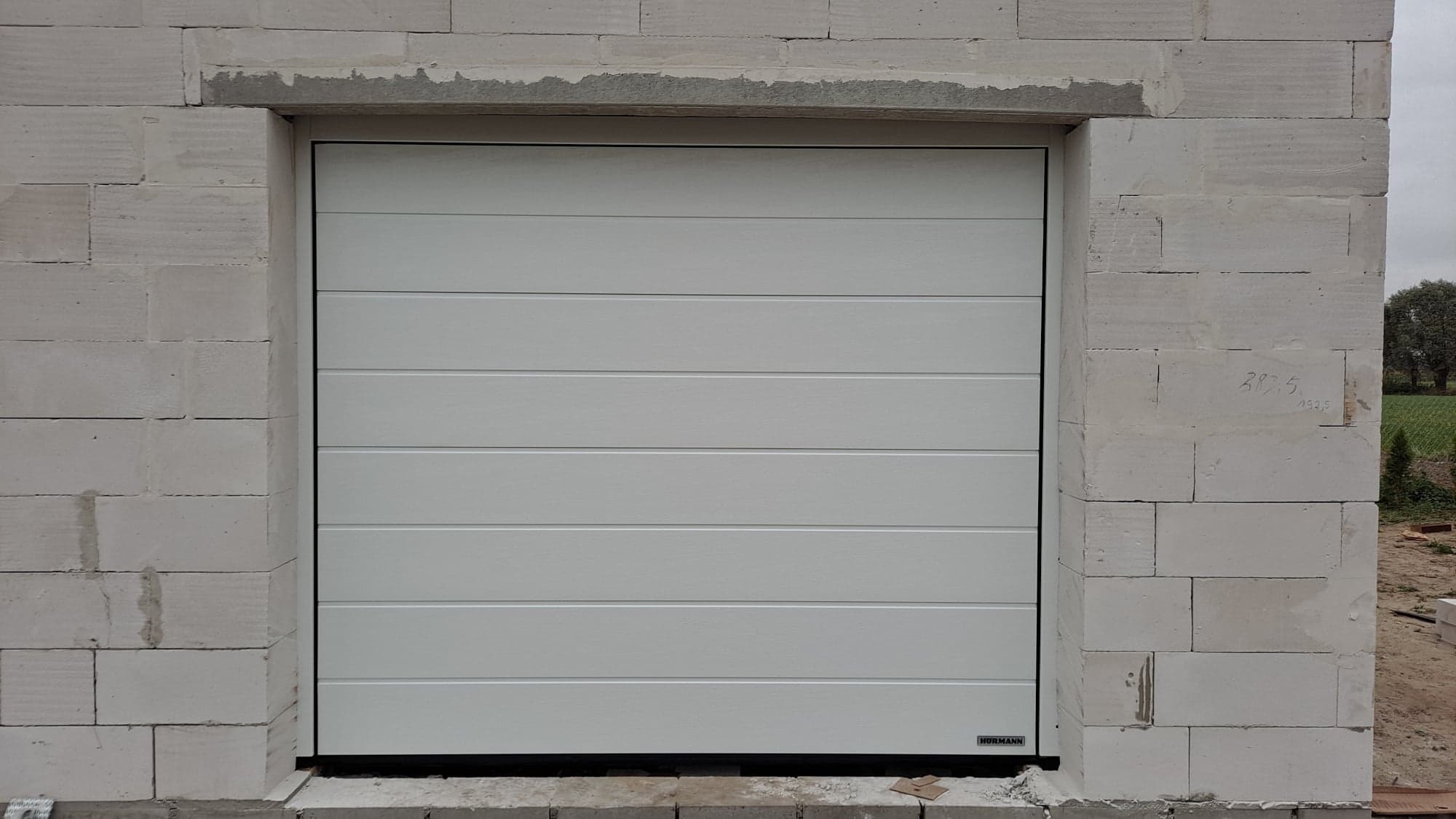 Biała segmentowa brama garażowa marki Hormann zamontowana w surowym murze z betonu komórkowego, widoczne niedokończone obramowanie otworu bramy.