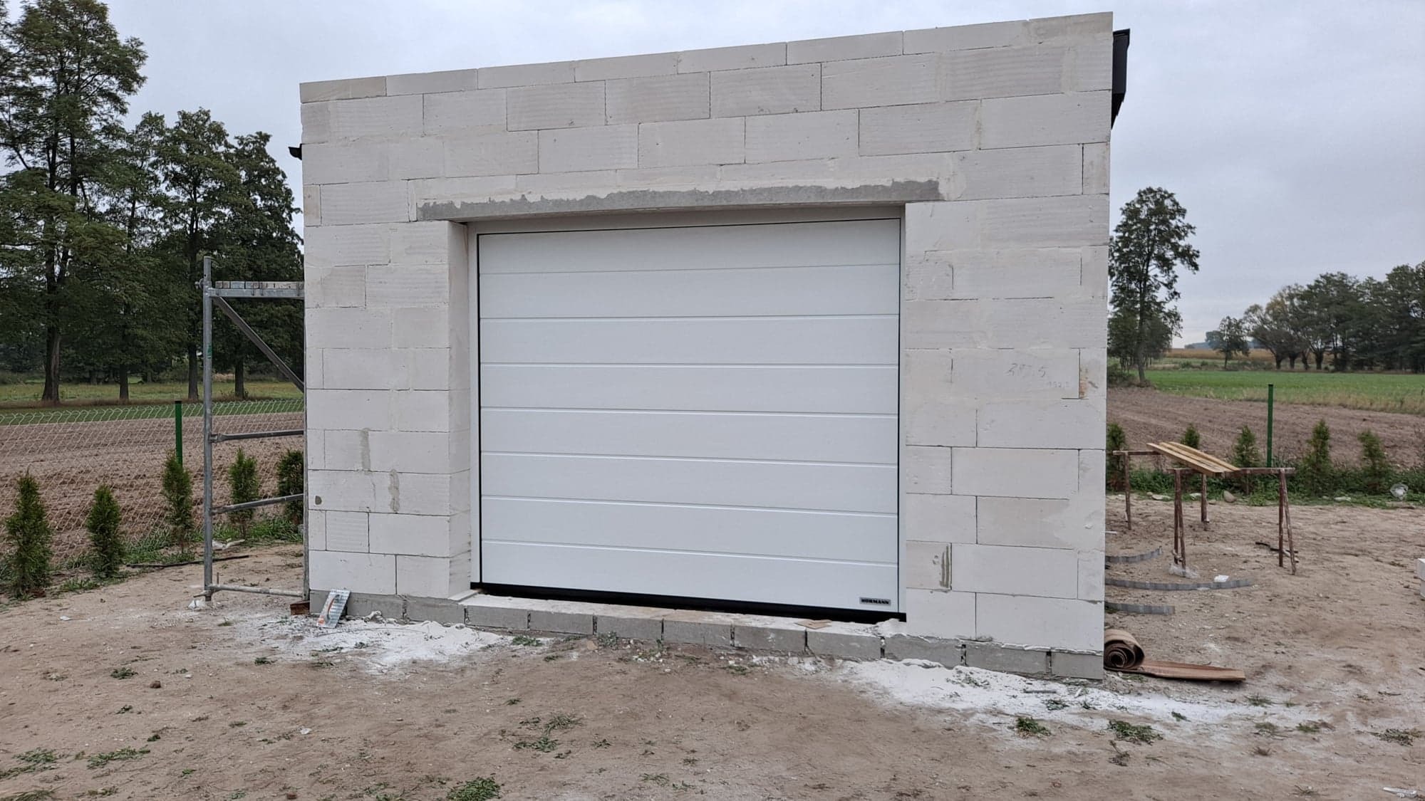 Biała segmentowa brama garażowa zamontowana w surowym murze z bloczków betonowych, widoczne rusztowanie i elementy wykończeniowe na zewnątrz.
