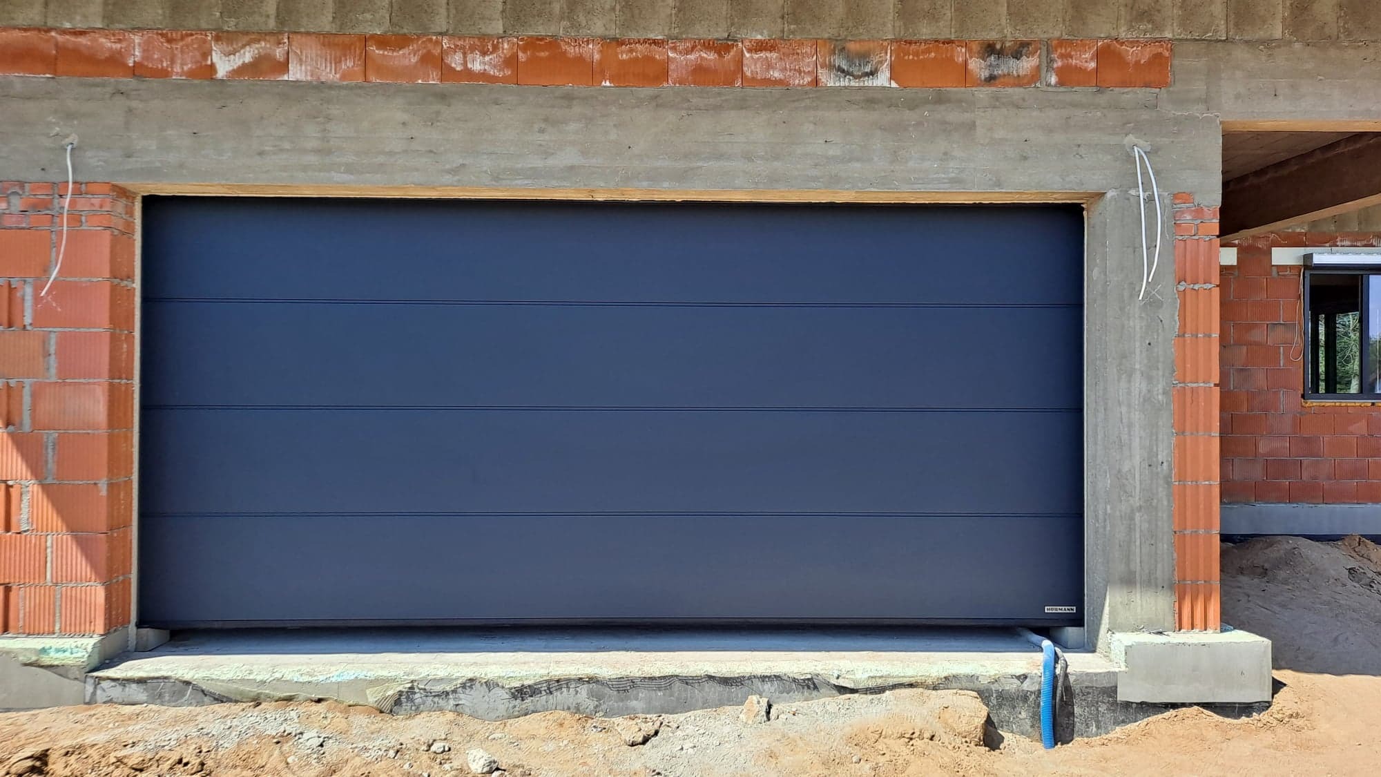 Zamknięta, pozioma, grafitowa brama garażowa segmentowa z drobnymi przetłoczeniami, zamontowana w surowym otworze budynku z cegły i betonu, widoczne niedokończone prace wykończeniowe.