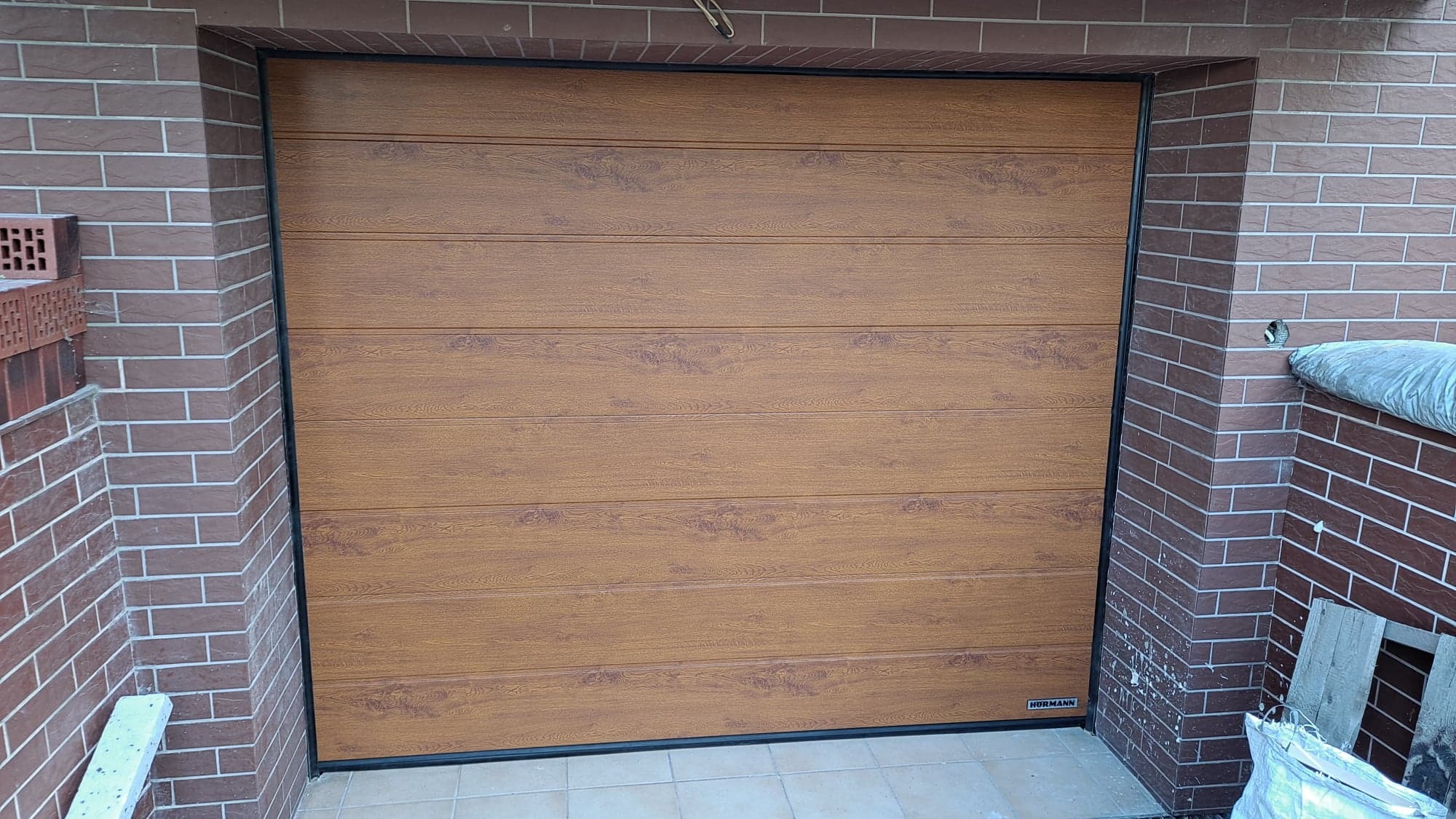 Zamknięta brama garażowa segmentowa, panelowa, o fakturze drewna, z czarną ramą, zamontowana w murze z brązowej cegły.