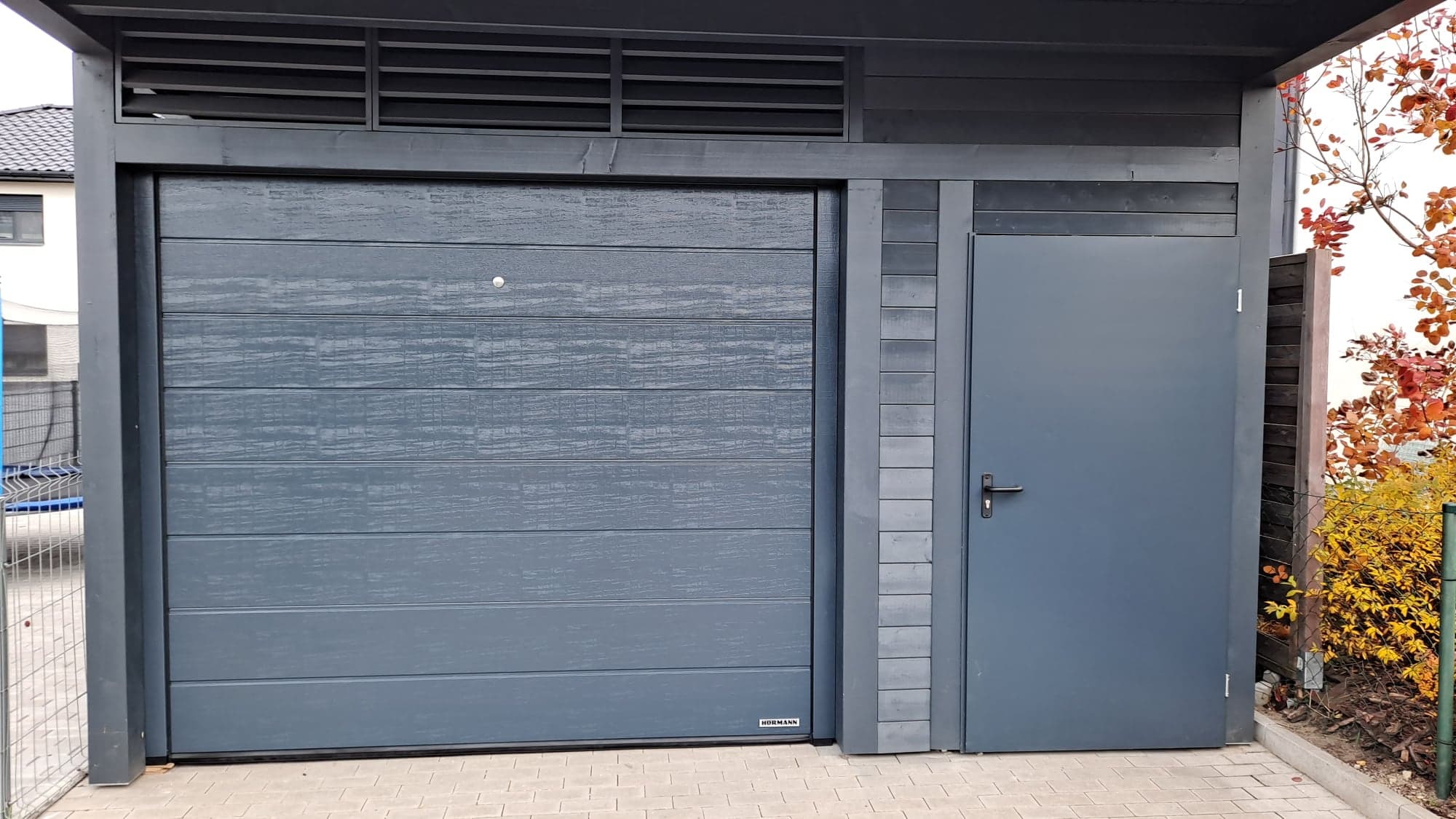 Szara brama garażowa segmentowa z poziomym wzorem desek, umieszczona w konstrukcji z szarego drewna, obok pojedyncze drzwi wejściowe, na zewnątrz kostka brukowa i jesienne krzewy.