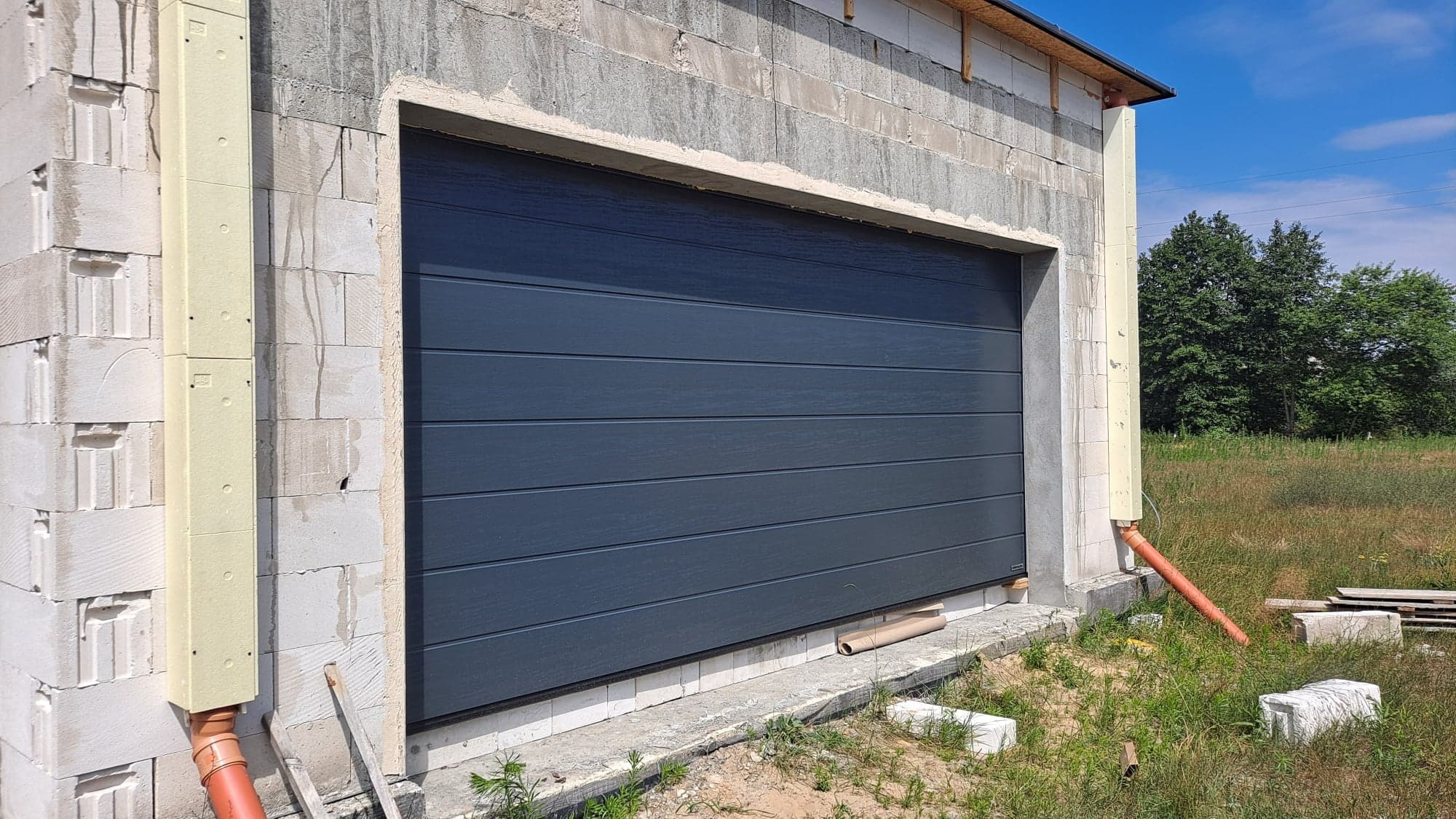 Szara segmentowa brama garażowa w trakcie montażu, widoczna na tle surowej ściany z bloczków betonowych i fragmentów ocieplenia, z rurą spustową po lewej stronie.