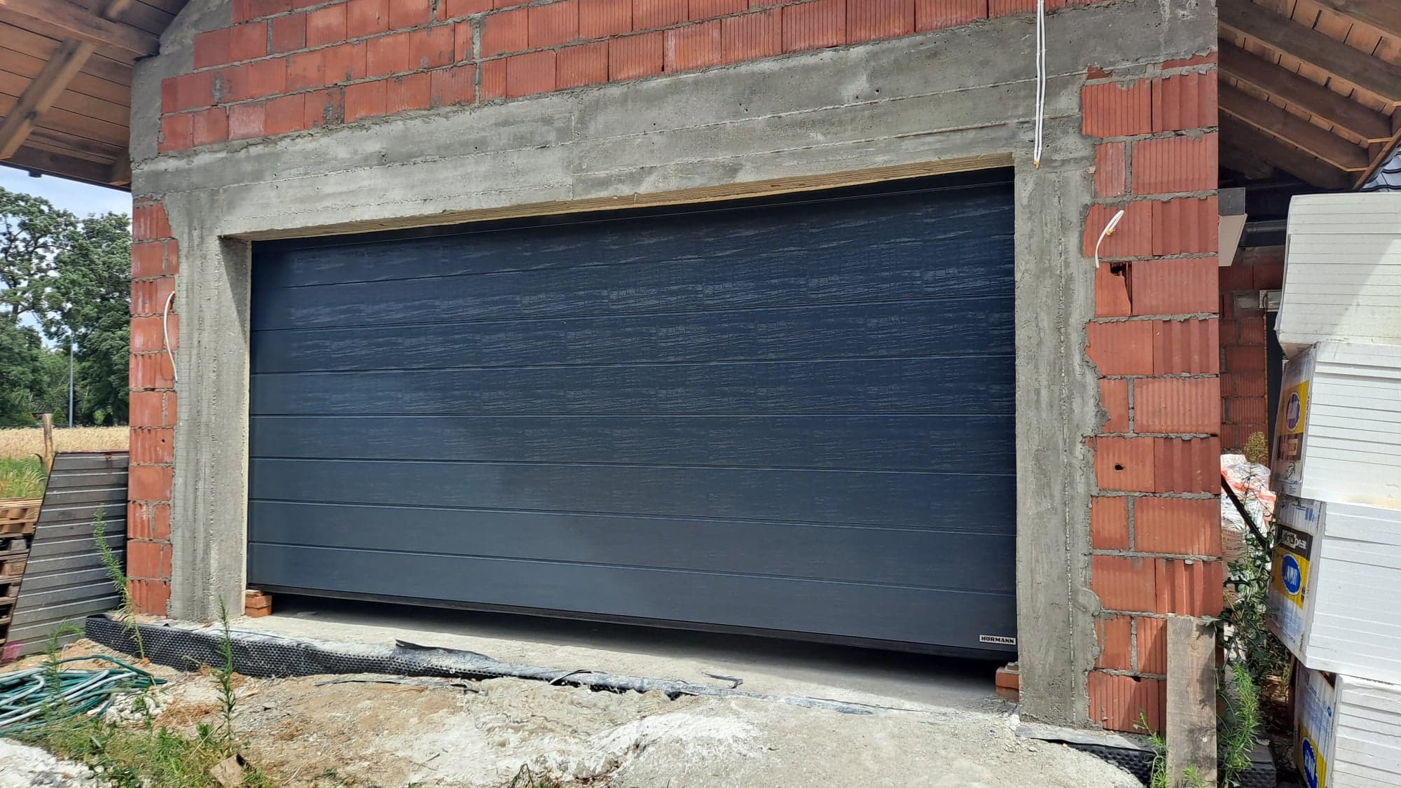 Szara segmentowa brama garażowa marki Hormann, zamontowana w surowym stanie budynku z czerwonej cegły i betonu, widoczne materiały budowlane obok.