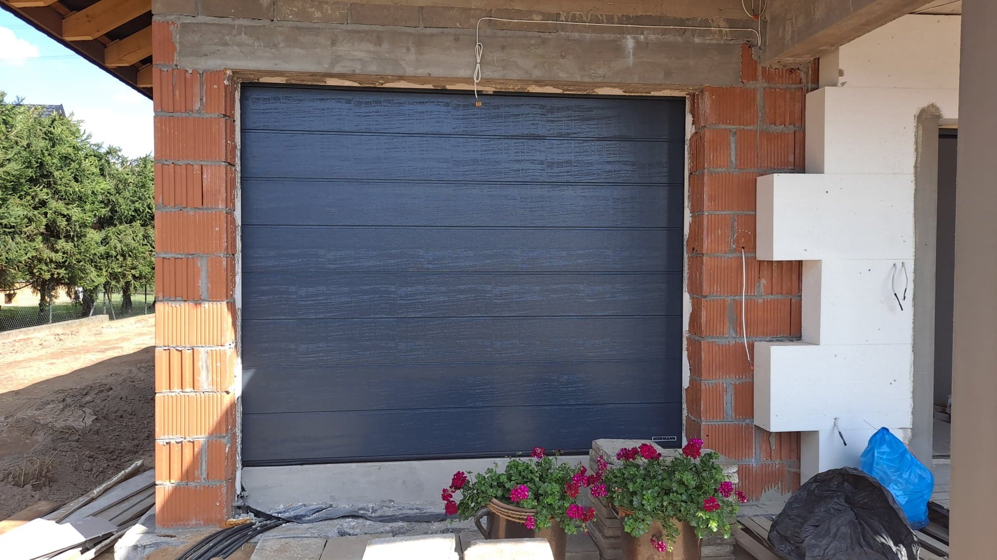 Nowa, antracytowa brama garażowa segmentowa z poziomymi przetłoczeniami, zamontowana w surowym otworze budynku z cegły kratówki i ociepleniem ze styropianu, z donicami z różowymi kwiatami u podstawy.