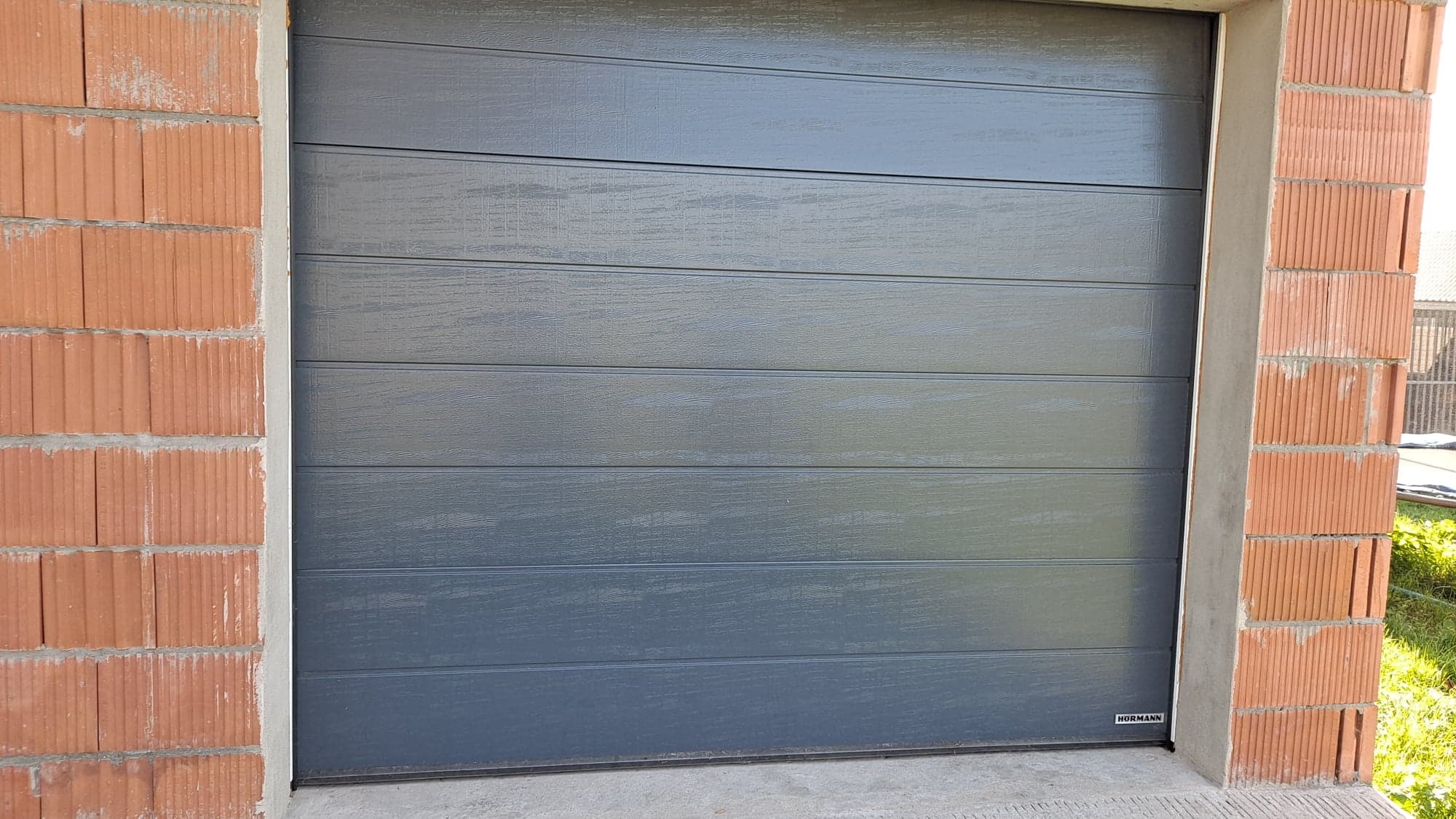 Zamknięta, segmentowa brama garażowa w odcieniach szarości, z poziomymi panelami o fakturze drewna, zamontowana w surowym otworze murowanym z cegły. Widoczny fragment logo Hormann w prawym dolnym...