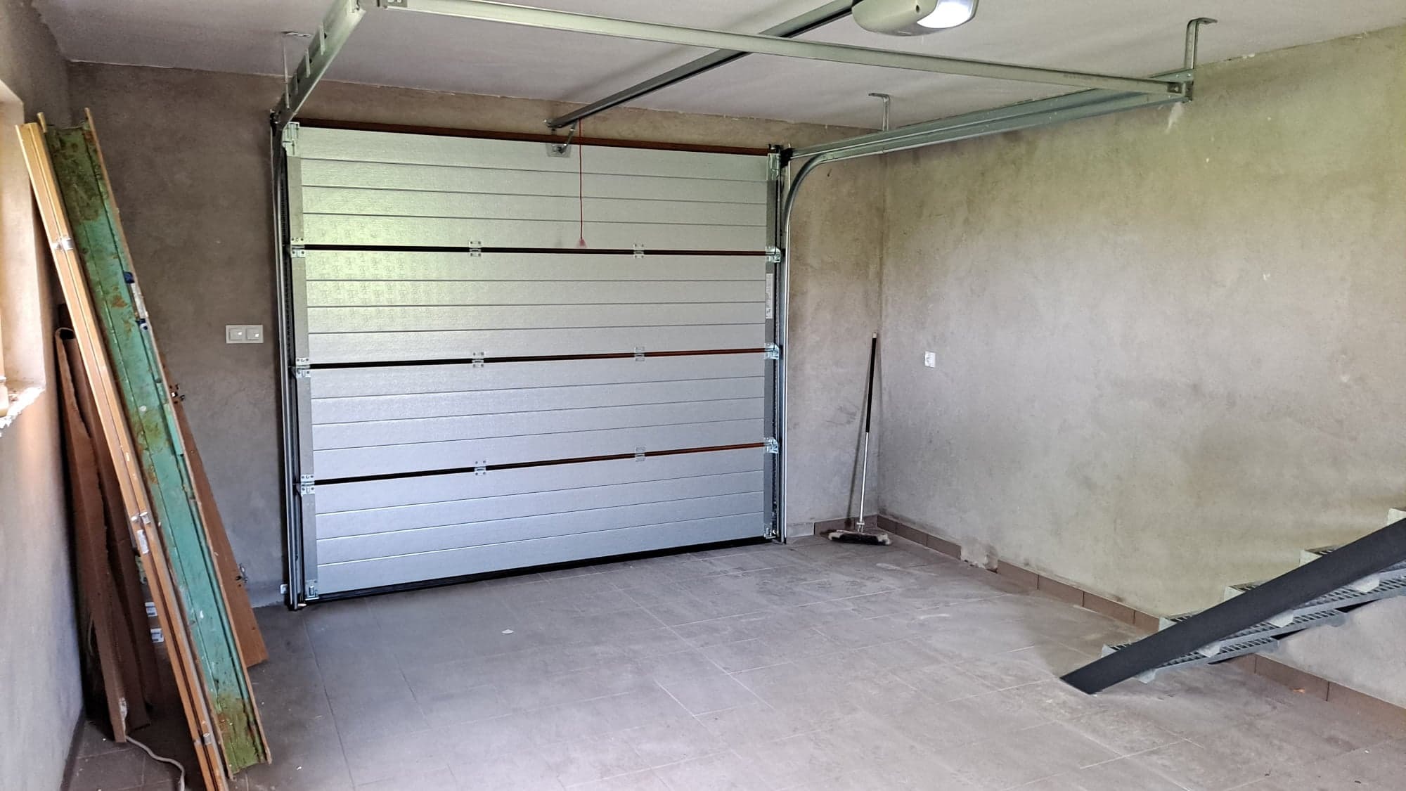 Wnętrze garażu z zamontowaną segmentową bramą garażową w kolorze srebrnym, widoczne prowadnice sufitowe, ściany w surowym tynku, pod ścianą oparta stara stolarka okienna.