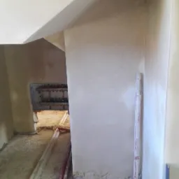 Świeżo nałożone, gładkie tynki na ścianach w trakcie remontu, widoczne rury instalacyjne na podłodze i fragment skrzynki rozdzielczej w głębi pomieszczenia.