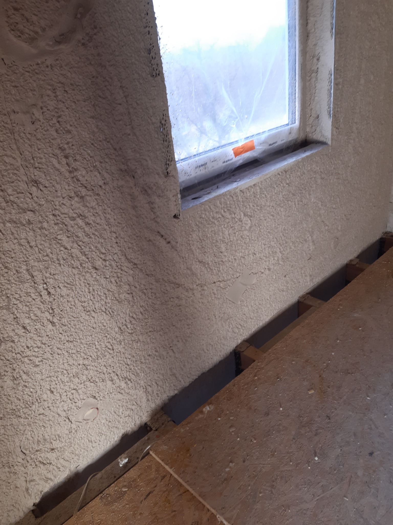 Świeżo nałożony, jasny tynk na ścianie wewnętrznej z widocznym oknem i fragmentem drewnianej podłogi w trakcie remontu.