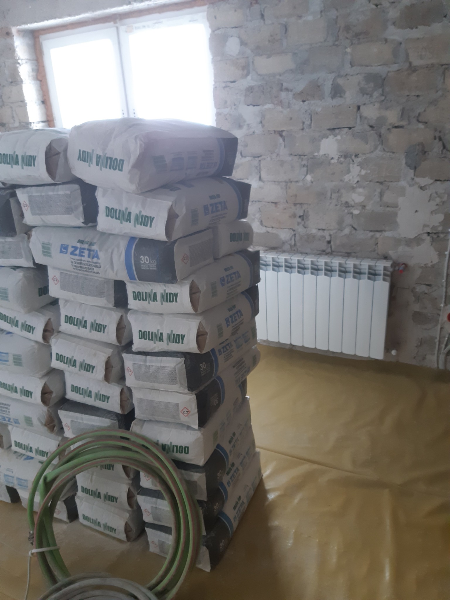 Stos worków z zaprawą gipsową Dolina Nidy Zeta 30kg na folii ochronnej, widoczny fragment ściany z surowej cegły i grzejnik.