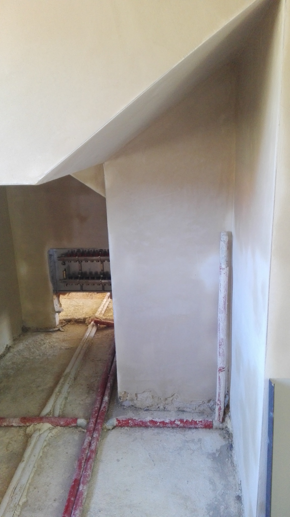 Świeżo nałożone, gładkie tynki na ścianach w trakcie remontu, widoczne rury instalacyjne na podłodze i fragment skrzynki rozdzielczej w głębi pomieszczenia.