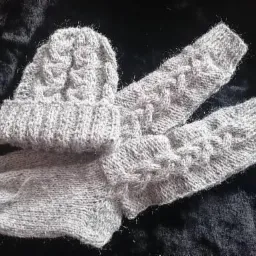 Ręcznie robiony komplet: szara czapka z warkoczowym wzorem i ocieplacze na ręce, leżące na ciemnym, teksturowanym tle.